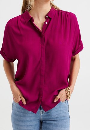 Femme portant un chemisier magenta à manches courtes avec des boutons et un jean bleu clair, mains dans les poches, et un bracelet épais en or au poignet droit.