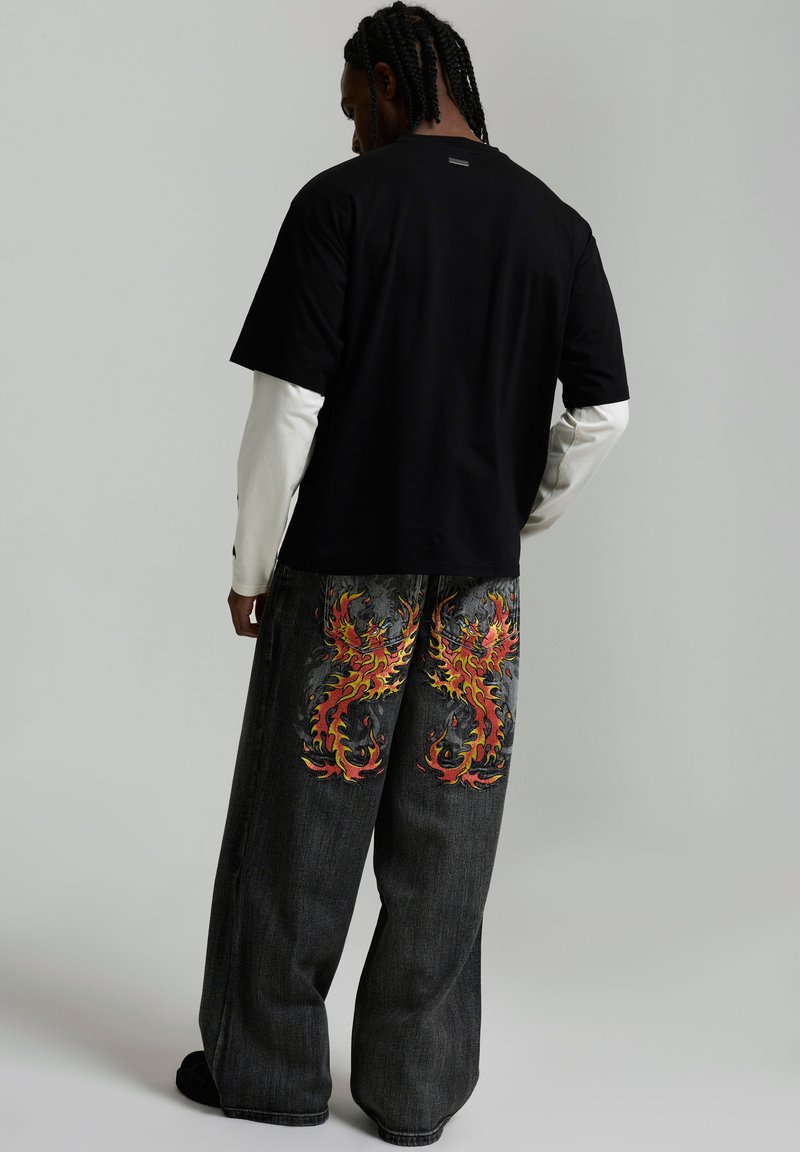 Person mit geflochtenem Haar, die ein schwarzes kurzärmeliges Shirt über einem weißen langärmeligen Shirt und weite schwarze Jeans mit roten und gelben Flammendesigns trägt.