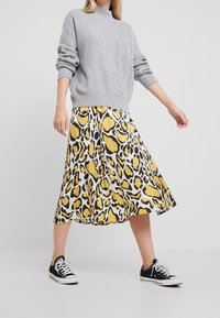 Šedý pletený svetr s vysokým výstřihem, který je zkombinován s volnou sukní se žluto-černým leopardím vzorem a černými teniskami.