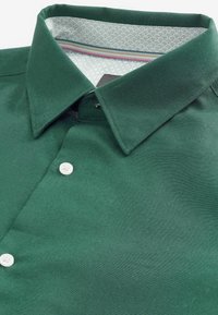 Cămașă verde cu guler clasic, nasturi albi și o căptușeală interioară cu model și accente subtile. Textură de țesătură netedă.