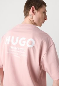 Light pink cotton t-shirt featuring a large white graphic with "HUGO," "MCMXCIII," and decorative text on the back. Classic cut with short sleeves.