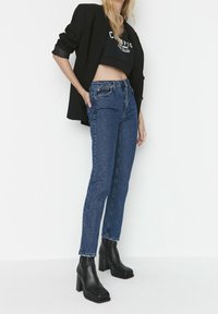 Jeans in denim blu a vita alta con una vestibilità dritta, abbinati a un top corto nero e stivaletti neri con tacco a blocco. Blazer nero indossato sulle spalle.
