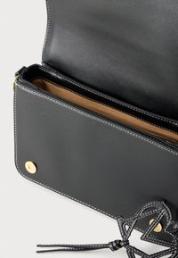 Μαύρη δερμάτινη τσάντα χειρός με ομαλή υφή, εσωτερικό σε αντίθετο μπεζ χρώμα, χρυσά μεταλλικά στοιχεία και διακοσμητική μαύρη λεπτομέρεια με κόμπους.