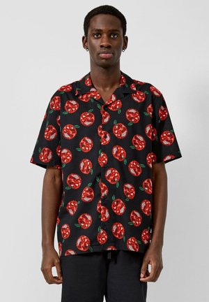 Mann trägt ein schwarzes kurzärmeliges Hemd mit Knöpfen und einem sich wiederholenden roten und weißen „Salty Fruits“-Blumenmuster, steht vor einem einfarbigen Hintergrund.