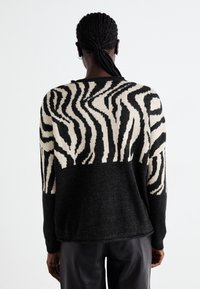 Personne avec des cheveux tressés portant un pantalon noir et un pull rayé zèbre noir et beige, vue de dos sur un fond blanc.