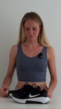 Zwarte sportschoenen met witte accenten, ronde neus en gestructureerde zool. Draag door een persoon in een grijze crop top, staand tegen een lichte achtergrond.