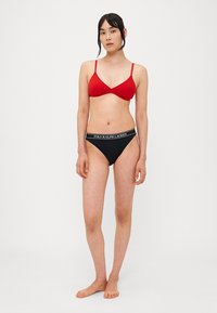 Sarkans trīsstūriski bikini augšdaļa ar plānām siksnām, apvienota ar melniem bikini apakšdaļām, kuru viduklī ir balts zīmola elastīgums. Gluda auduma, minimāla dizaina.