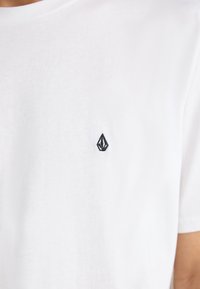 White cotton T-shirt with a crew neck and short sleeves. Features a small black embroidered logo on the left chest area.