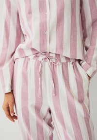 Femme portant une chemise à boutons rayée verticale rose et blanche et un pantalon à cordon assorti, une main dans la poche.