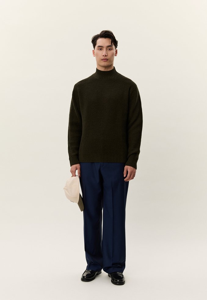 Egy férfi áll, sötétzöld kötött pulóvert, tengerészkék nadrágot és fekete cipőt visel, bal kezében bézs színű sapkát tart, egyszínű háttér előtt.