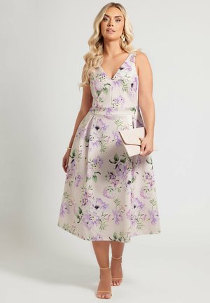 Kukallinen midi-mekko, jossa on V-pääntie ja leveät olkaimet. Mekossa on vaalea beige pohja ja purppuran värisiä kukkia, istuva bodice ja levenevä hame.