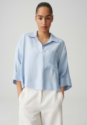 Vrouw met haar naar achteren gebonden, draagt een lichtblauw overhemd met kraag en zak, en witte hooggetailleerde broek, staand met handen in de zakken.