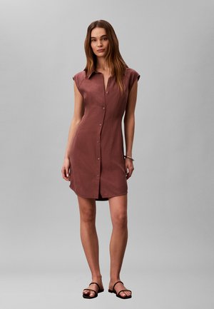 Kobieta stojąca w brązowej sukience bez rękawów z guzikami z przodu i czarnych sandałach na jednolitym szarym tle.