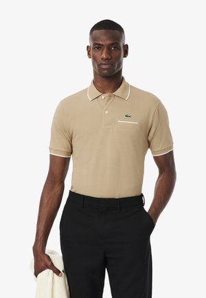 Polo beige avec des accents blancs, manches courtes et un logo vert. Associé à un pantalon noir et tenant un pull couleur crème.