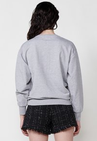 Felpa grigia in cotone con maniche lunghe, scollo rotondo e polsini a costine, abbinata a pantaloni corti neri con texture, orlo sfilacciato e motivo a quadri.