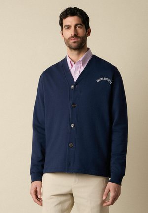 Uomo che indossa un cardigan blu navy con bottoni e il logo "Brooks Brothers" sopra una camicia a righe rosa e pantaloni beige, in piedi contro uno sfondo semplice.