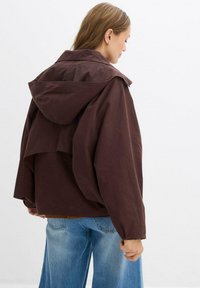 Donna che indossa un giubbotto oversized marrone scuro con cappuccio e maniche ampie, abbinato a jeans azzurri, in piedi davanti a uno sfondo bianco uniforme.
