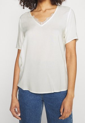 Vrouw die een wit T-shirt met korte mouwen en V-hals met kanten randje draagt, gecombineerd met blauwe spijkerbroek, staand tegen een effen achtergrond.