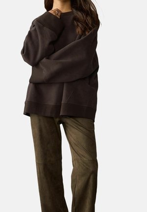 Femme portant un sweat-shirt marron foncé oversize et un pantalon en daim vert olive, main près du menton, sur fond uni.