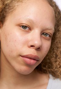 Ritratto di una persona con capelli biondi ricci, pelle chiara, trucco facciale minimo e un colore labbra naturale e sottile. Tessitura della pelle dettagliata visibile.