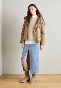 Veste brune matelassée avec capuche, pull crème à côtes, jupe en denim bleu fendue sur le côté, et bottines beiges. Textures douces et style décontracté.