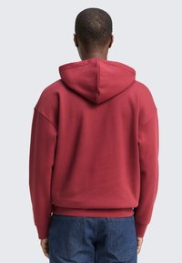 Bordeauxroter Kapuzenpullover mit großer, hinterer Kapuze, oversized Passform und gerippten Bündchen. Der Stoff wirkt weich, mit einer glatten Textur und minimalen Designdetails.