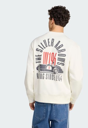 Mężczyzna w białej bluzie z grafiką vintage samochodu wyścigowego i napisem „The Silver Arrows W196 Racing Since 1954” na plecach.