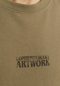 Camiseta de algodón verde oliva con un diseño de texto impreso en negro que dice "OBRA DE ARTE CONTEMPORÁNEA" en la parte del pecho.