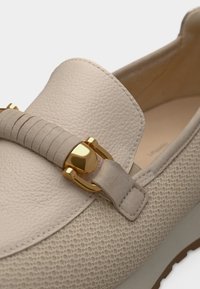 Φλατ μοκασίνια σε ανοιχτό μπεζ χρώμα με υφή, που διαθέτουν διακοσμητικό στοιχείο σε χρυσό τόνο και λεπτομέρεια με λείο δερμάτινο λουράκι. Σχεδιασμός με επίπεδη σόλα.