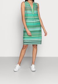 Kvinna i ärmlös, knälång grön mönstrad klänning med V-ringning och vita sneakers, stående mot en enfärgad grå bakgrund.