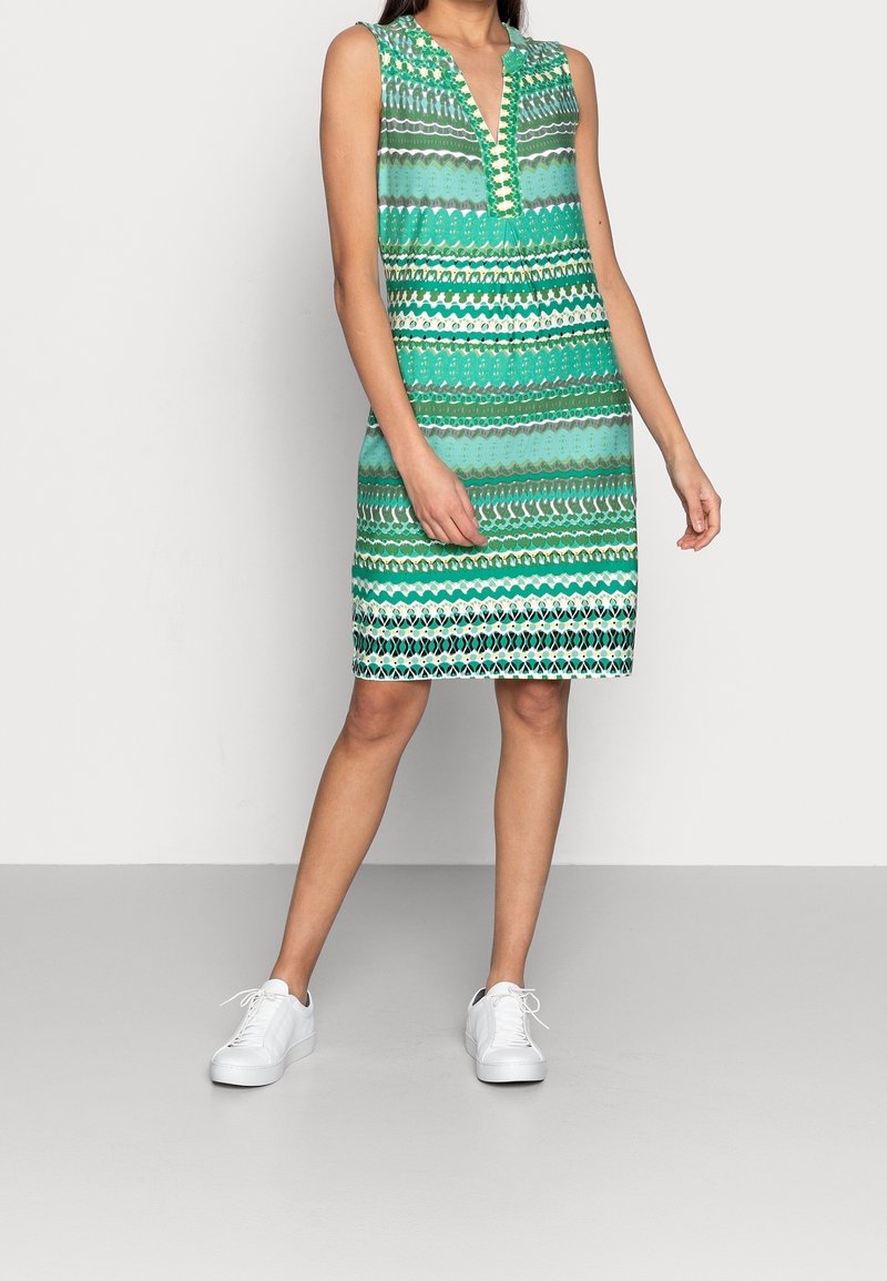 Kvinna i ärmlös, knälång grön mönstrad klänning med V-ringning och vita sneakers, stående mot en enfärgad grå bakgrund.