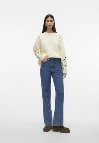 Sudadera crema con bordado floral en las mangas, combinada con vaqueros de talle alto en azul y zapatos gruesos de color marrón oscuro. Fondo blanco liso.