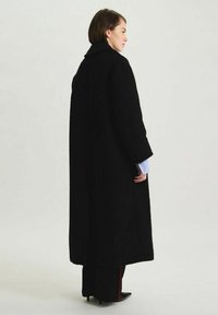 Manteau long noir en tissu texturé, avec un large col et une coupe droite. Présente des manches amples et un design minimaliste.