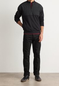Sudadera negra con cuello con cremallera, panel superior texturizado y sección inferior lisa, combinada con pantalones negros y zapatos oscuros.