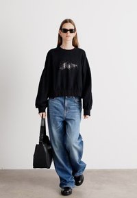 Felpa nera oversize con design stampato, jeans a gamba larga blu, borsa nera e scarpe chunky nere. Gli accessori includono occhiali da sole neri.