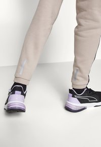 Czarne buty sportowe z fioletowymi akcentami, zestawione z beżowymi dresami z czarnymi paskami po bokach i odblaskowymi detalami na ściągaczach.