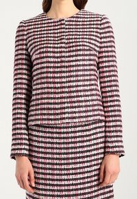 Veste en tweed tissé rose, noire et blanche avec un col rond, des manches longues et une fermeture à boutons, présentant un motif à carreaux texturé.