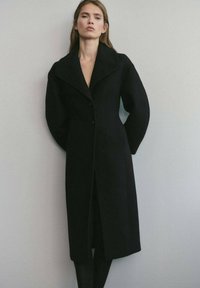 Abrigo de lana negro con un corte entallado, gran cuello y cierre de botones. Presenta mangas voluminosas y un diseño elegante y minimalista.