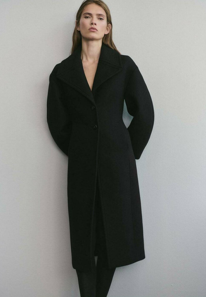 Abrigo de lana negro con un corte entallado, gran cuello y cierre de botones. Presenta mangas voluminosas y un diseño elegante y minimalista.