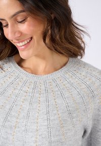 Suéter de punto gris con un cuello acanalado y tachuelas doradas decorativas dispuestas en líneas verticales a lo largo del cuerpo. Textura suave.