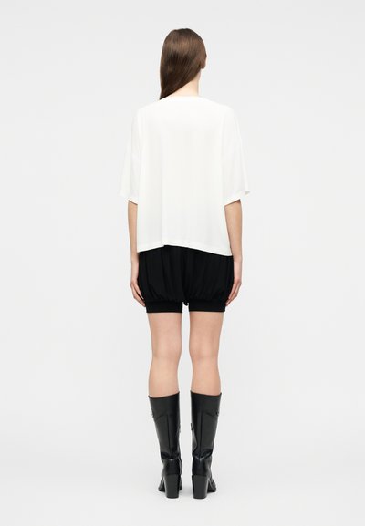Γυναίκα που στέκεται με την πλάτη στραμμένη, φορώντας άνετο λευκό τοπ, μαύρα φουσκωτά σορτς και μαύρες ψηλές μπότες με τακούνι μέχρι το γόνατο, σε απλό φόντο.