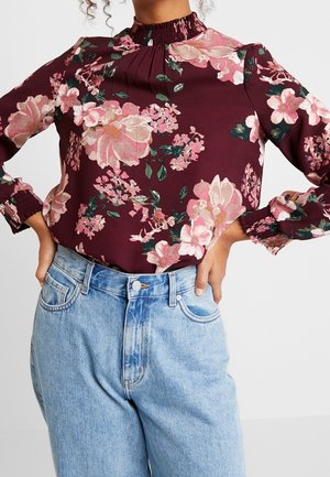 Kobieta w bordowej bluzce w kwiaty z długim rękawem, marszczonym wysokim kołnierzem oraz jasnoniebieskich, wysokich dżinsach, z rękami opartymi na biodrach.
