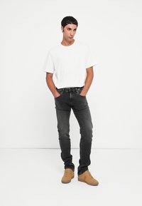 White short-sleeve T-shirt, black slim-fit jeans, and beige sneakers. Model poses with hands in pockets against a plain, light background.