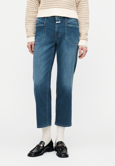 Femme portant un jean bleu raccourci, un pull en maille crème, des chaussettes blanches et des mocassins en cuir noir à boucles dorées.