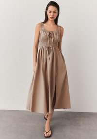 Ein beiges Maxikleid mit einem gerafften Oberteil, breiten Trägern und einem Kordelzug, ausgestattet mit einem fließenden Rock und leichtem Stoff.