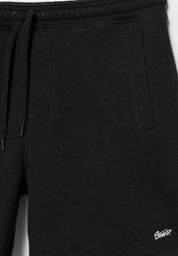 Čierne tepláky s elastickým pásom, šnúrkou na stiahnutie, bočnými vreckami a malým bielym vyšívaným logom "Santa" blízko ľavého vrecka.