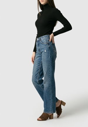 Woman wearing a black long-sleeve turtleneck, high-waisted blue jeans, and brown fringed heeled sandals, standing with one hand on her hip.