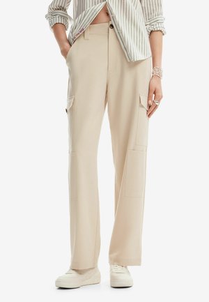 Beżowe szerokie spodnie typu cargo noszone z białymi trampkami, zestawione z koszulą w paski z podwiniętymi rękawami oraz srebrną biżuterią na prawej ręce.