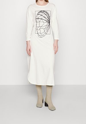 Mujer con un vestido largo blanco con un diseño abstracto de cara negra en la parte delantera, calcetines beige de punto y zapatos negros con tacón, de pie sobre un suelo gris.