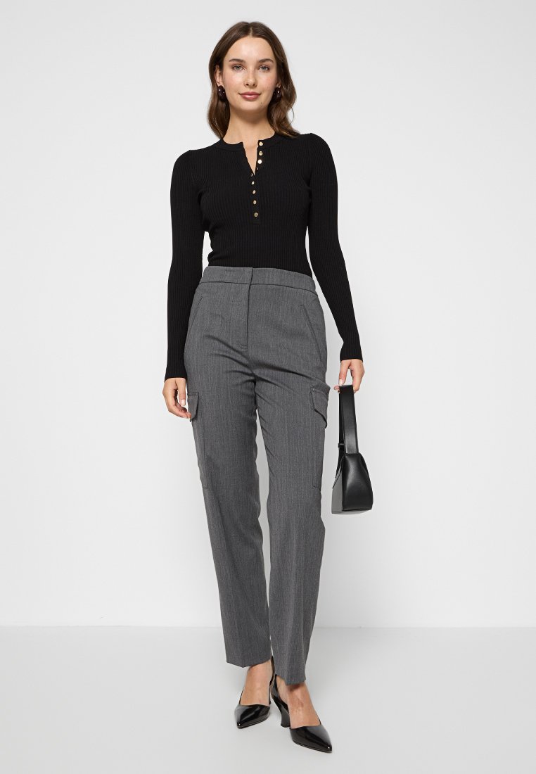 Schwarzes, geripptes Langarm-Oberteil mit Henley-Ausschnitt und goldenen Knöpfen, kombiniert mit grauen, gestreiften Hosen und schwarzen spitzen Pumps.